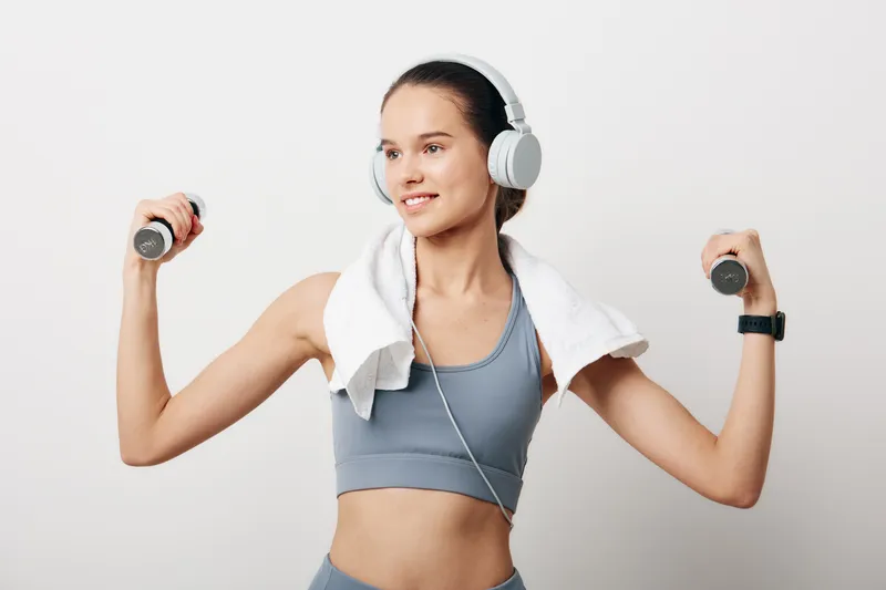 Woman fitness enthusiast lifts dumbbells in a studio, wearing headphones with a towel around the neck, performing a strong workout with focus, showcasing sport and an active lifestyle.