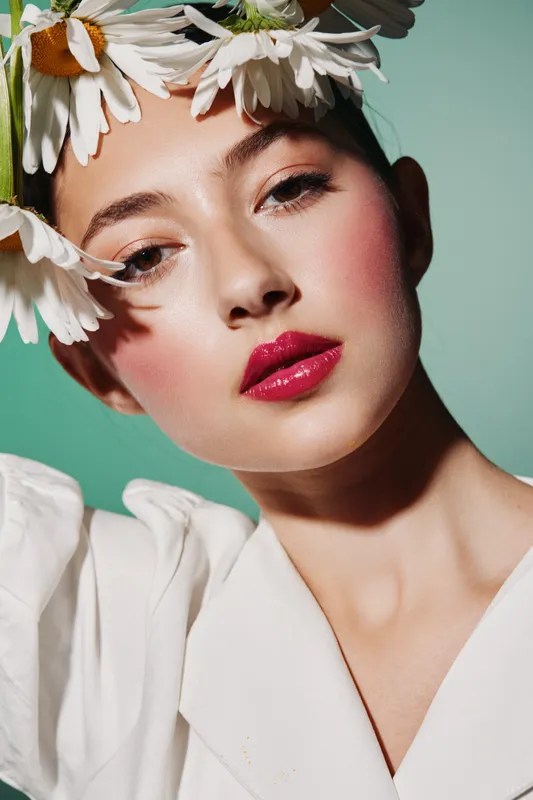 Close-up portrait of a young woman with glossy red lips and blunt bangs wearing a flower crown, dressed in white against soft green background. Beauty fashion concept.