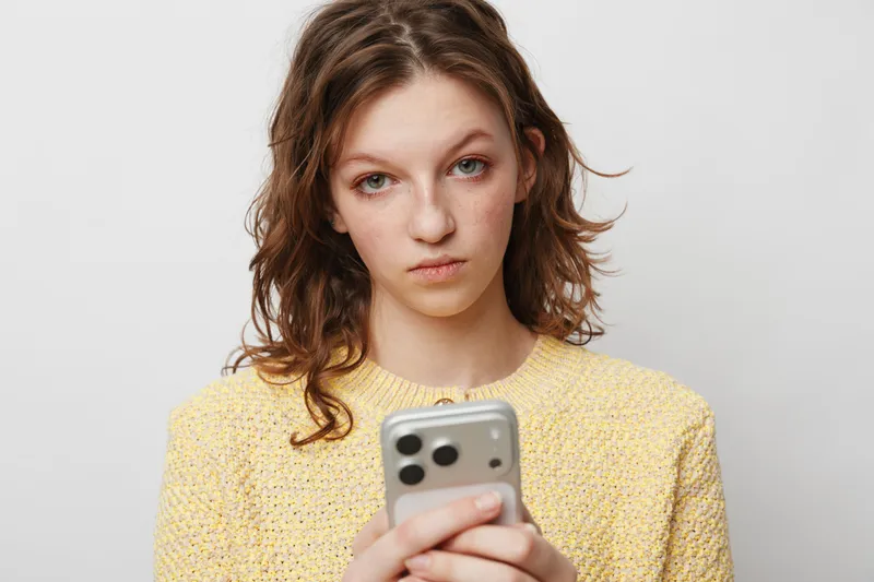 Young woman in yellow sweater looks serious while holding smartphone in hands. Isolated on white background. Concept of communication, technology, and emotion.
