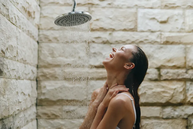 Woman enjoying outdoor shower with water flowing, standing against textured stone wall, wet hair, relaxed mood in natural light setting.