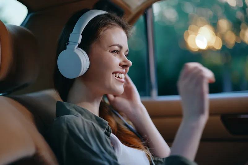 Young woman enjoying music with headphones in car, smiling with casual outfit, warm bokeh background, capturing joyful emotions and comfort.