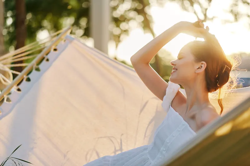 Beautiful young woman relaxing in a hammock, enjoying the sunshine, wearing a white summer dress, exuding happiness and tranquility, in a serene outdoor setting
