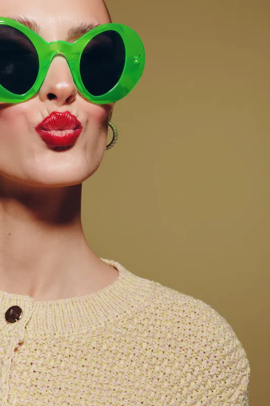Close-up of woman wearing large green sunglasses and red lipstick making a kiss face. She wears a beige knitted sweater against a plain beige background. Fashion and beauty concept.