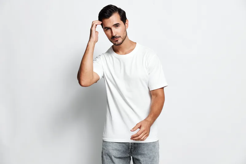 Handsome young man in a white t-shirt stands against a light grey background, looking thoughtful while touching his hair and engaging with the camera. Style concept.