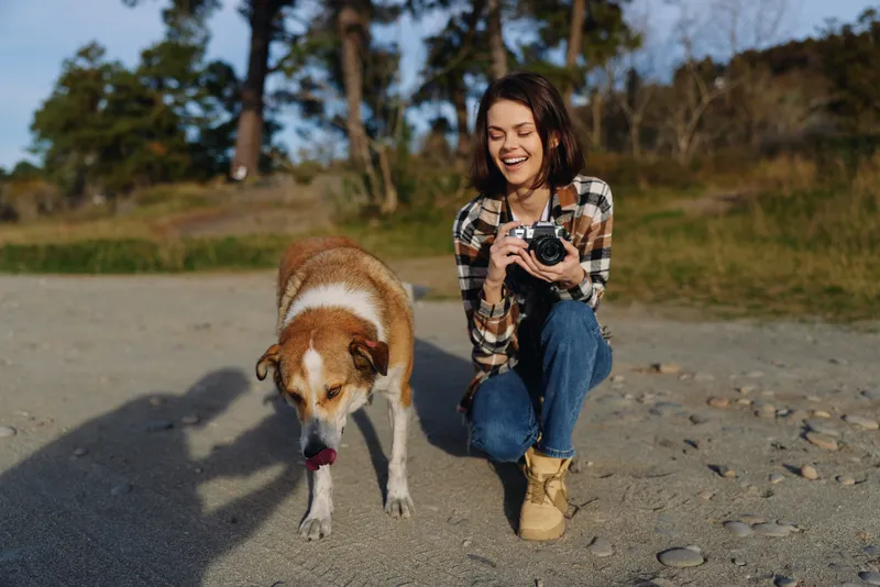 Female photographer crouches beside a dog on a sunny beach, capturing candid outdoor moments with a camera, celebrating nature, playfulness, and companionship in bright daylight