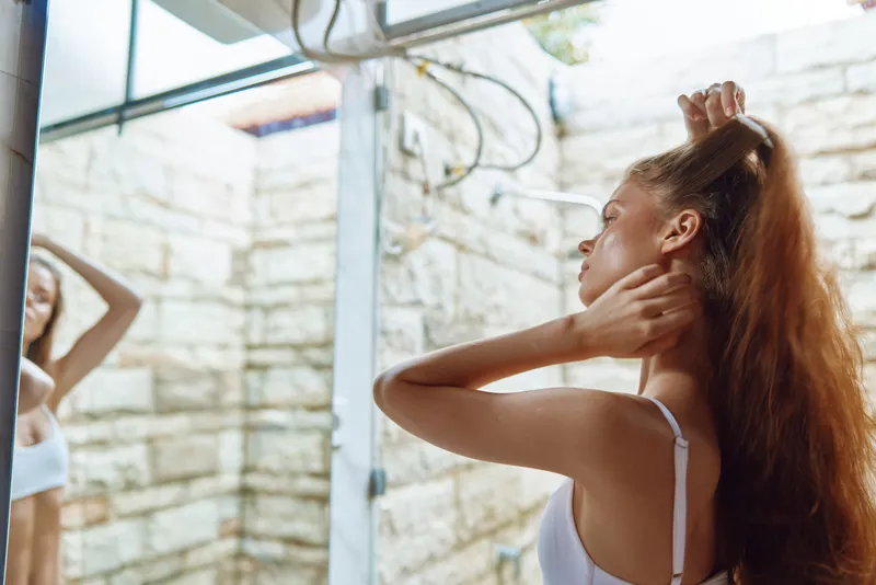 young woman with long red hair adjusting it in front of a mirror in natural light, wearing white tank top, indoors with stone wall background