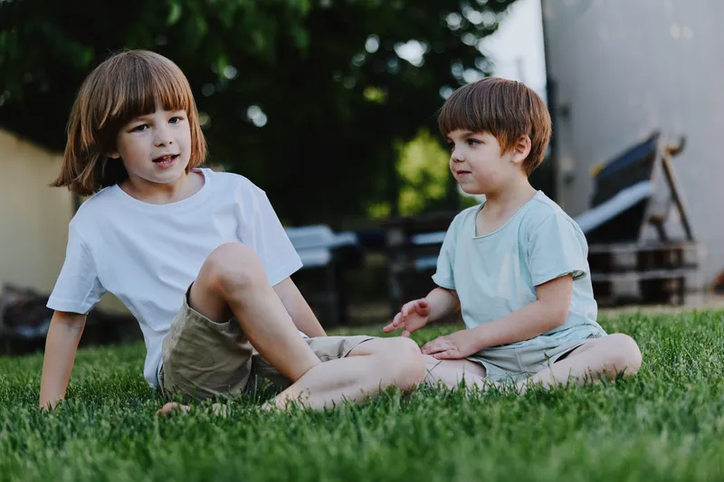 Two boys sitting on grass, enjoying a sunny day. They are playing and interacting, showcasing the joy of childhood and friendship in a casual outdoor setting.