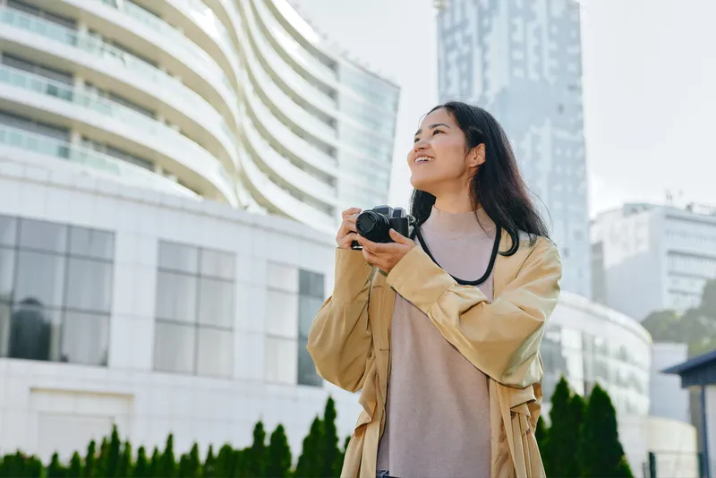 Woman with camera in an outdoor urban setting, capturing skyline and architecture, casual jacket, sunlit day, candid street scene, travel mood and creative photography moment in city life