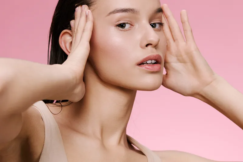 Young woman with fresh skin and neutral makeup, holding her face against a soft pink background, conveying calm and beauty