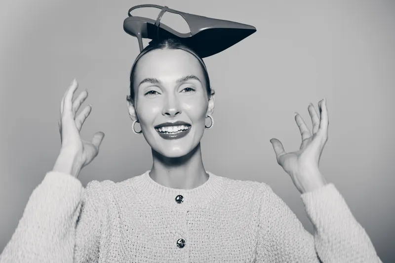 Smiling white woman with light skin wearing textured sweater and unique black headwear gestures with hands in studio. Concept for fashion and creativity.