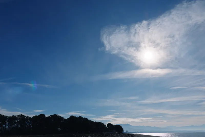 Landscape of clear sky and sun over calm water, with a distant tree line along the coast and wispy clouds, capturing peaceful outdoor scenery for nature and travel projects