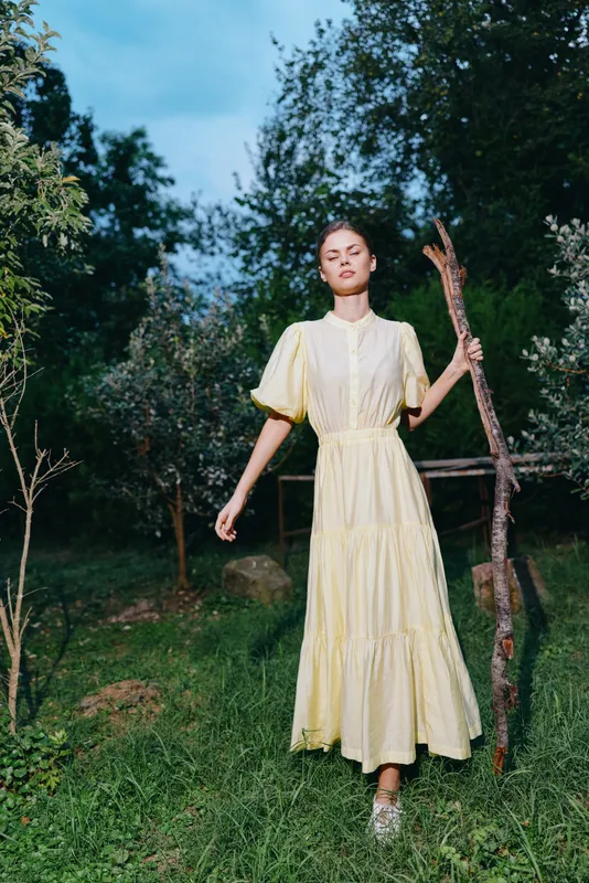 Woman in yellow dress standing in meadow holding stick, outdoor portrait with natural light, authenticity inclusive serene