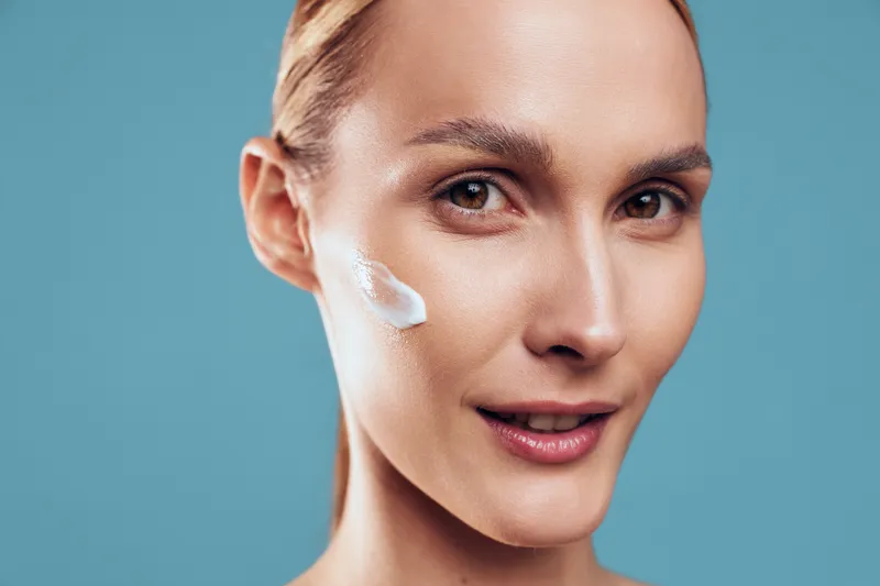 Woman, face, cream, skincare, beauty, closeup, selfcare, wellness, studio, portrait of woman applying white face cream on cheek, soft light, blue background, skincare routine
