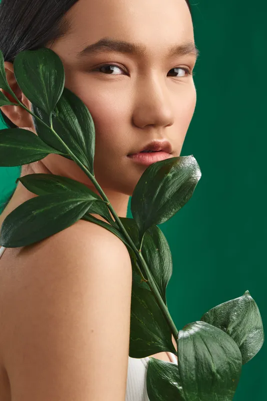 Asian woman gazes softly toward the camera, framed by lush green leaves. The natural contrast highlights calm beauty and presence, creating a serene, modern aesthetic for lifestyle and wellness