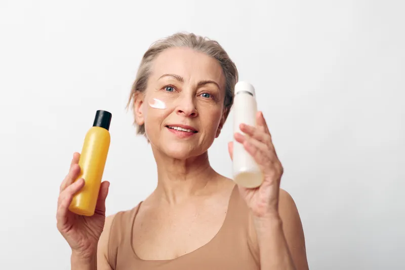 Mature woman with short hair applies cream on face holding two cosmetic bottles on white background. Concept of skincare, beauty routine, anti aging and self care for healthy skin.