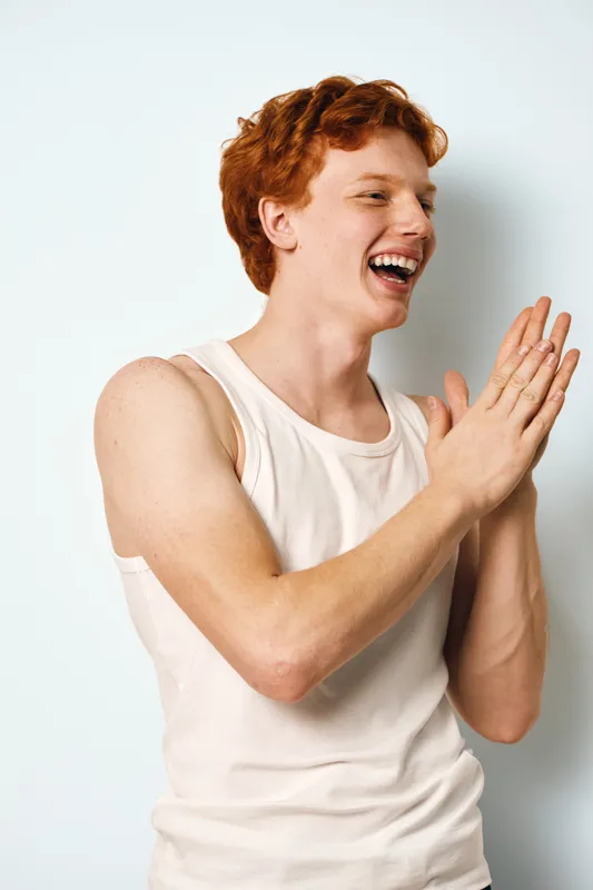 Young man with red hair and freckles laughing and clapping hands wearing sleeveless white shirt against plain background, casual expression of joy and happiness in studio portrait.