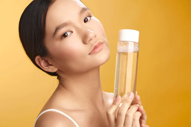 Asian woman holds a clear bottle against a warm yellow backdrop, presenting a calm, poised expression that conveys beauty and wellness, a simple yet elegant product moment.