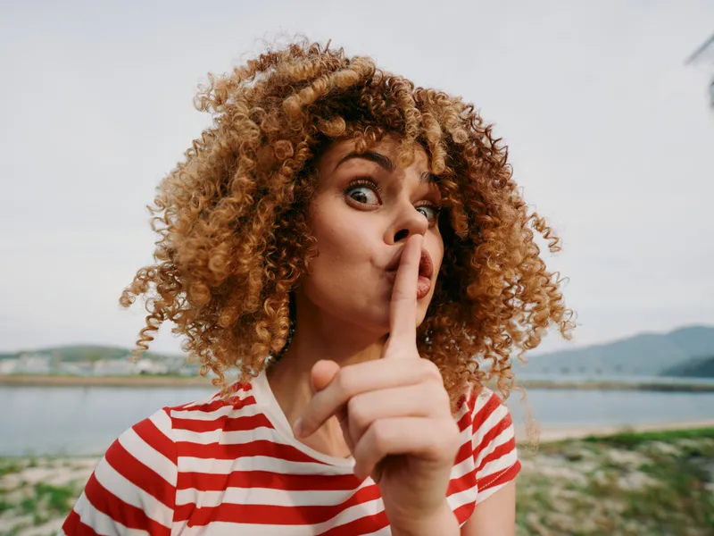 Portrait of a young woman with curly hair making a shushing gesture, finger to lips, outdoors by a lake and hills, wearing a red and white striped tee.