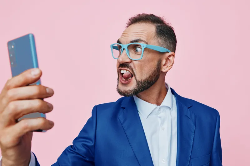 A business professional man in a blue suit takes a dynamic selfie against a solid pink background, showing confident enthusiasm and bold, energetic emotion.