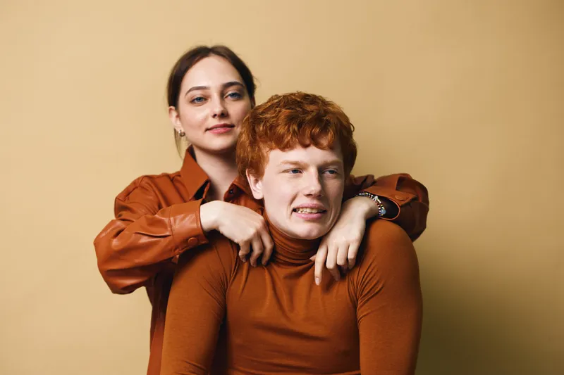 Young woman and man with red hair in brown clothing posing together against beige background with casual and friendly expression in studio portrait. Siblings or friends.