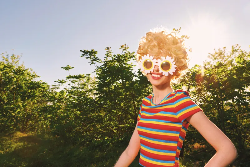 A cheerful woman in a striped shirt wears playful sunflower sunglasses in a sunlit park. Retro film color tones enhance the carefree, casual lifestyle vibe and outdoor portrait concept.