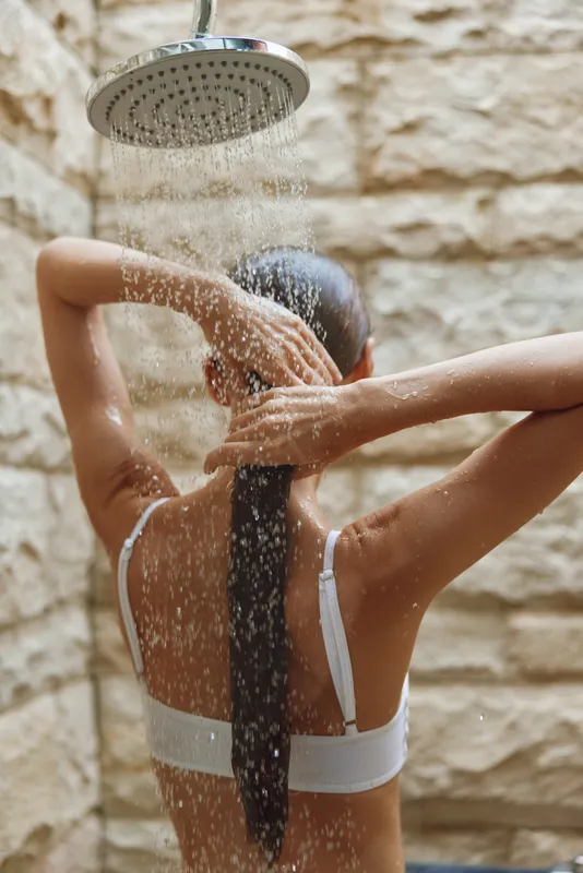 woman showering outdoors with long wet hair under rainfall showerhead, natural stone wall background, relaxed and refreshing moment in casual white underwear