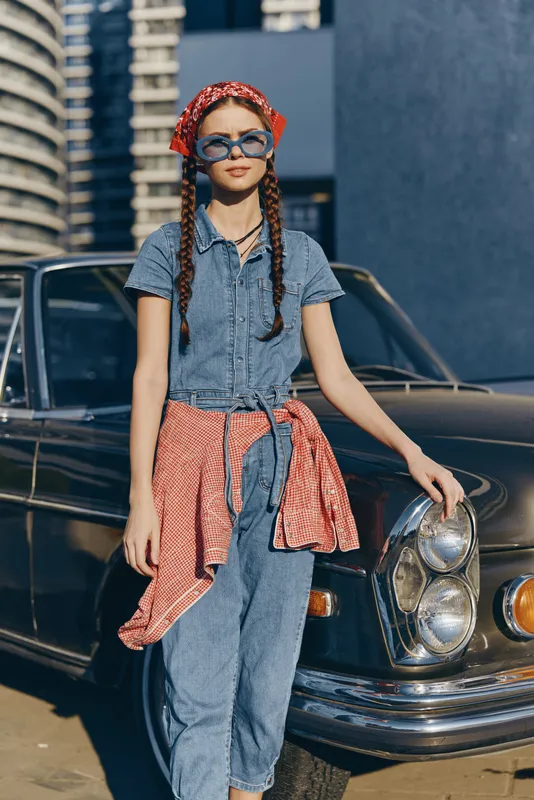 Modern boho-western denim street style woman with braided hair and sunglasses poses confidently against vintage car in warm film color tones.