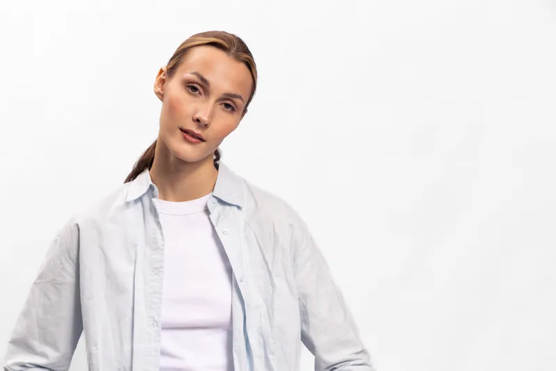 Young woman with serious expression wearing light shirt over white top stands against white background. Concept of casual fashion, confidence, and natural beauty in studio portrait.