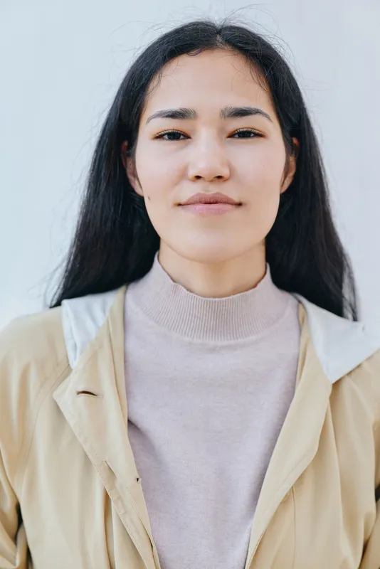 Calm woman with straight dark hair wearing a beige jacket over a light sweater against a pale wall, creating a clean minimalist fashion and lifestyle look for editorial use