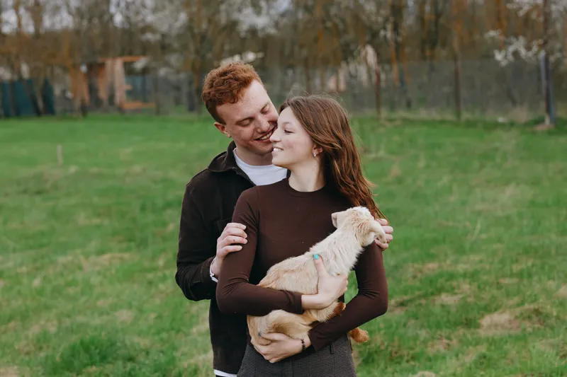 Happy couple outdoors with a small dog cuddling in a green park, man embraces woman as they smile, warm autumn light and casual style create a candid lifestyle portrait, showcasing affection and