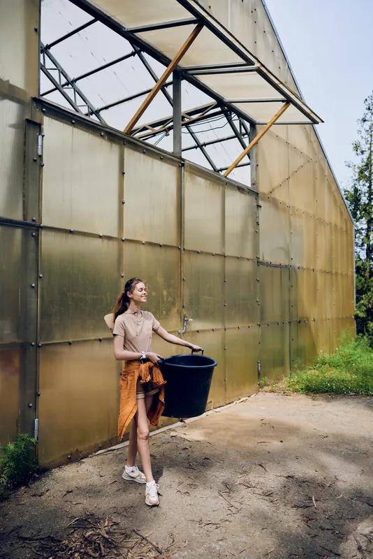 Woman in casual clothes walking outside greenhouse carrying a black bucket, gardening activity with smiling face and outdoor horticulture work near polycarbonate panels and greenery.
