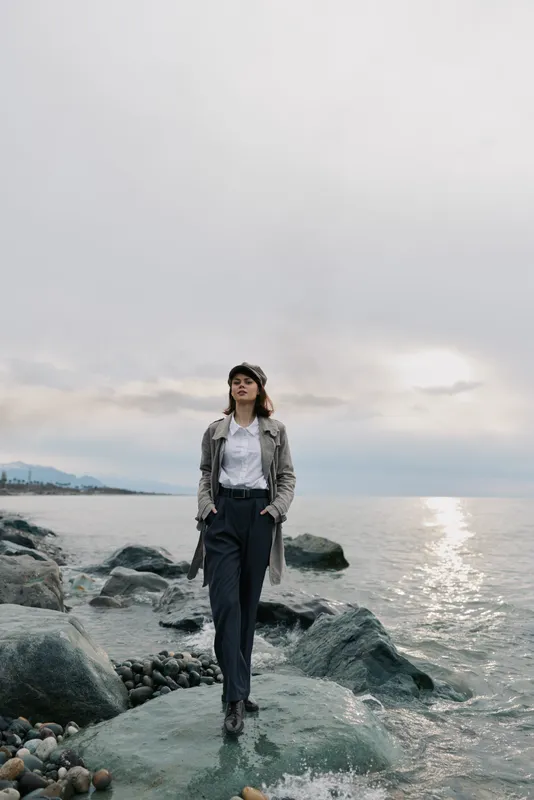 Young woman in stylish autumn outfit stands on rocky shore by calm sea under cloudy sky. Fashion portrait with natural light and muted colors for outdoor, travel, and seasonal concept.