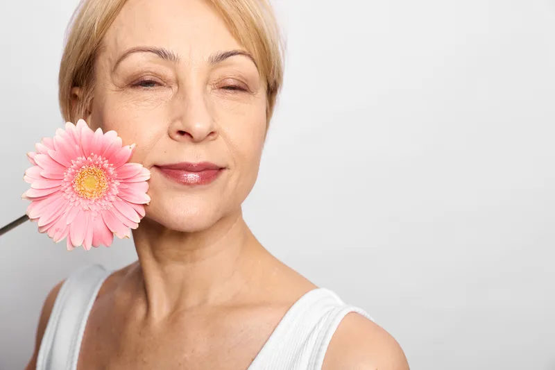 Woman closeup portrait, pink flower beside cheek, smiling softly, studio white background, gentle soft light, calm beauty mood, spring garden feeling