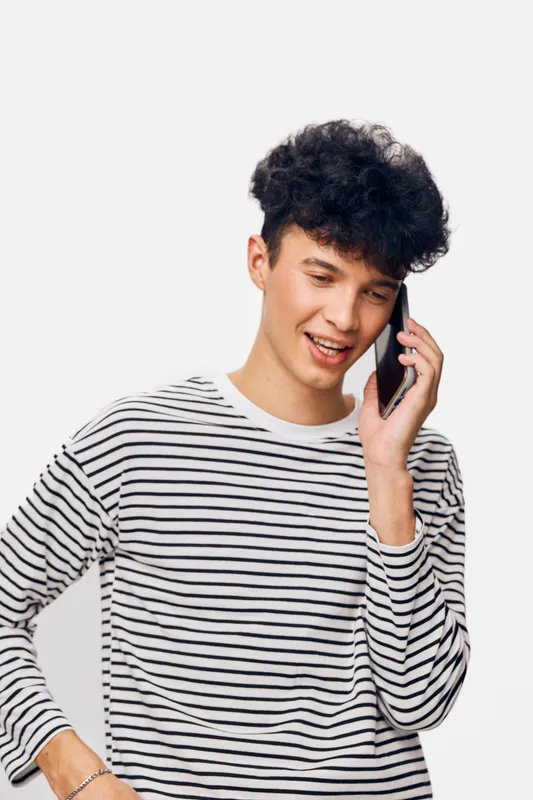 Young man talking on smartphone smiling and dressed in casual striped long sleeve shirt with curly hair against white background, cheerful expression, modern lifestyle.