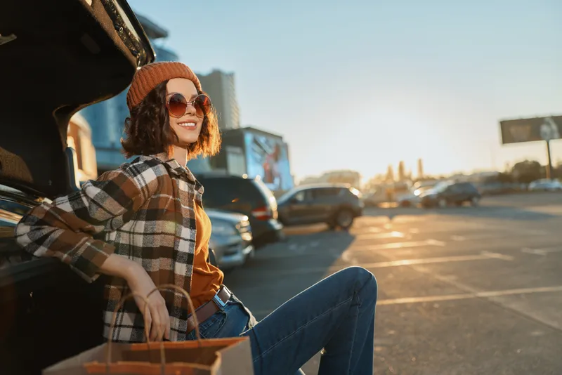 Woman in car trunk smiling with sunglasses and beanie in parking lot at golden hour, candid lifestyle scene highlighting authenticity, mindful living and emotional storytelling.