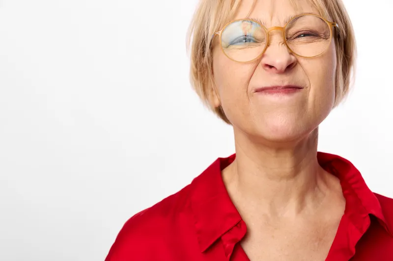 Closeup portrait of an elderly woman with short blond hair, wearing glasses and a red shirt, calm thoughtful expression, studio white background, soft light