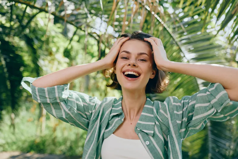 Joyful woman outdoors with hands in her hair, bright smile, lush greenery, and striped green shirt, capturing a carefree, sunny moment.