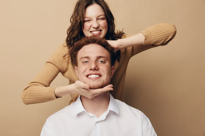 Happy young woman playfully holds smiling man face with hands. Casual couple in warm clothes poses on beige background. Concept of friendship, joy, and positive emotions together.