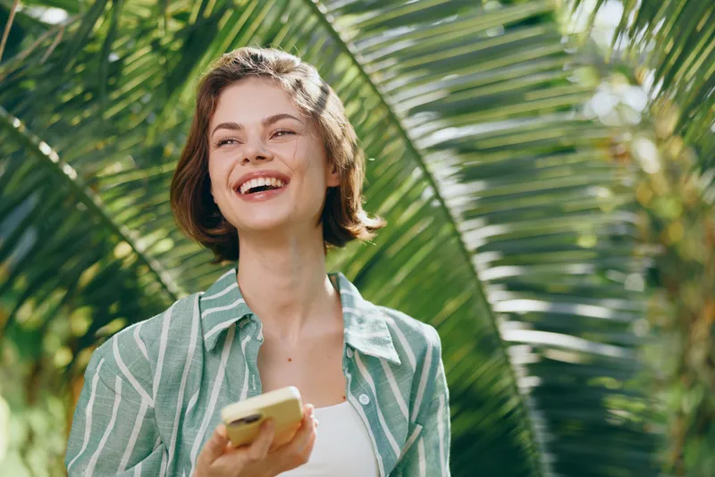 Cheerful young woman stands outdoors among tropical palm leaves, smiling while holding a smartphone.