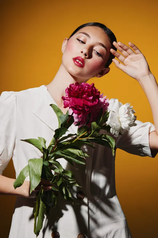Young woman with glossy red lipstick poses with vibrant pink and white flowers wearing a stylish white blazer, against a bright yellow background. Fashion beauty concept.