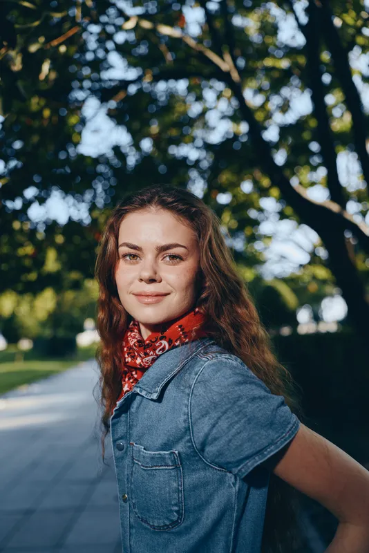 Lifestyle woman in modern boho-western denim street style smiling outdoors with warm film color tones and casual red scarf accessory during sunny day.