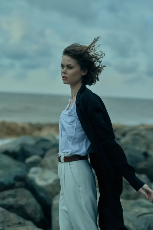 Woman on windy coastline wearing a jacket and light dress, standing among rocks with ocean backdrop, fashion forward mood and movement in moody coastal scenery