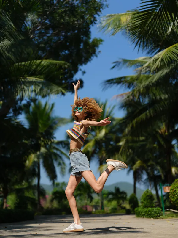 joyful young woman with curly hair jumping outdoors, wearing sunglasses and casual summer clothes, surrounded by tall palm trees and clear blue sky