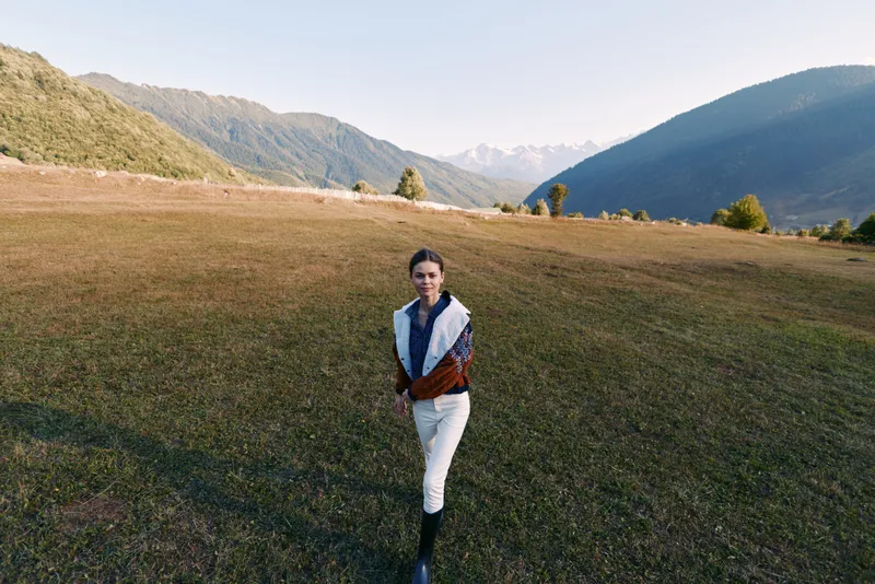 Woman standing in meadow in mountains valley outdoors, portrait of a solo traveler in open field and nature. Scenic green slopes and distant peaks create calm adventure atmosphere.