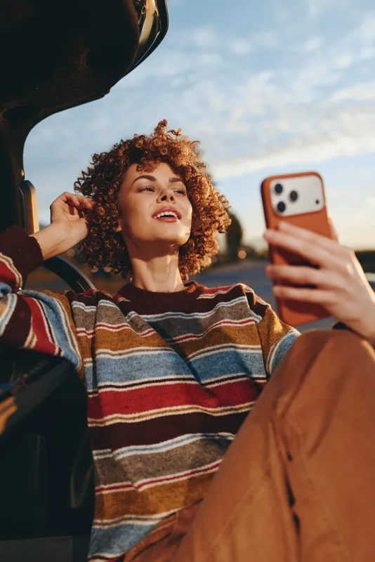 Woman lifestyle smartphone selfie curly hair smile friends outdoors autumn, friends adjust a hat, she smiles while taking a selfie, warm striped sweater, natural light candid portrait