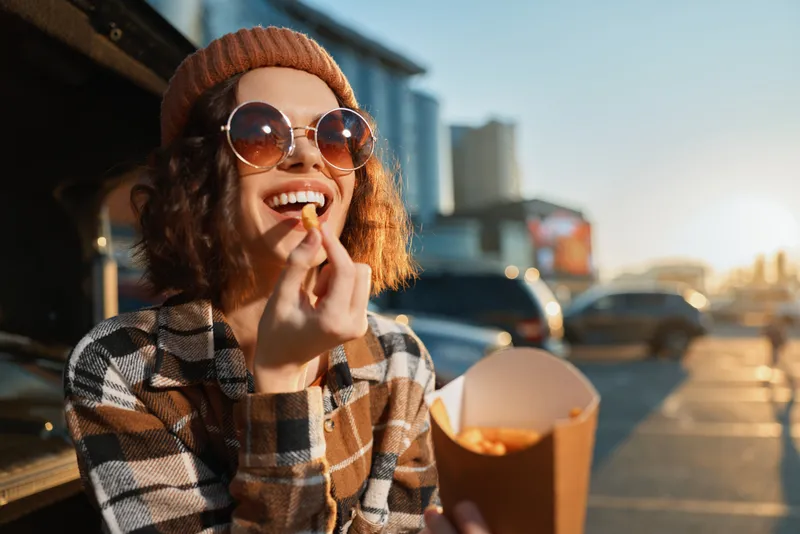 woman eating fries from takeout by car trunk at sunset, sunglasses and beanie, genuine smile in a candid lifestyle scene with golden hour glow, authenticity, mindful living and emotional storytelling