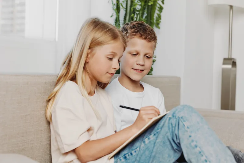 Two children sitting on a sofa drawing together with a pencil in a bright modern living room. Concept of creativity, childhood, learning, and friendship.