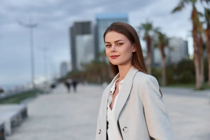Confident businesswoman in a stylish blazer, walking along a seaside promenade, exuding professionalism and elegance amidst a modern cityscape.
