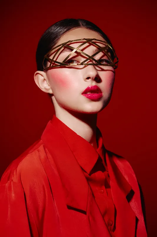 Creative beauty shot of a young woman with avant-garde eyewear and bold red makeup. She wears a red coat and shirt, posing against a matching vivid red background.