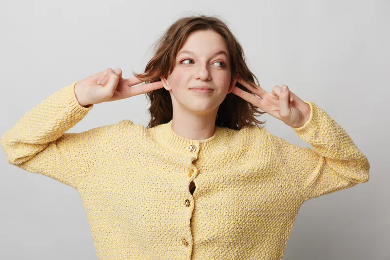 Young woman in yellow sweater covers ears with fingers and looks away with playful expression. Studio portrait on grey background. Concept of ignoring noise and playful mood.